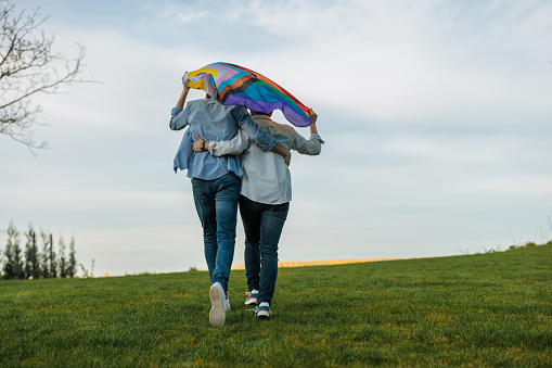 A Caucasian young adult gay couple is hugging and walking outside with a progress pride flag away from the camera.