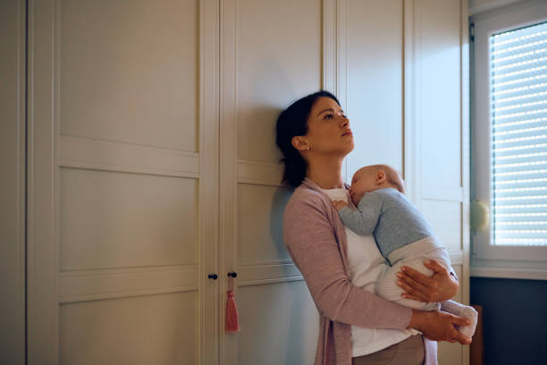 Young sad mother holding her sleeping baby in her arms at home. Distraught mother thinking of something while her baby is napping in her arms. Copy space. postpartum depression stock pictures, royalty-free photos & images