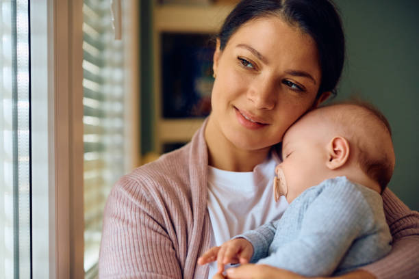 istock Smiling mother looking through the window while her baby is napping in her arms. 1489251313