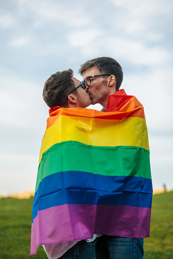 A happy gay emotional couple is outdoors caped with a pride flag and kissing.