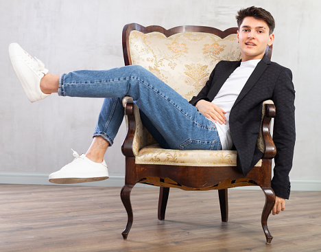 Smiling young guy teenager in business clothes posing while sitting in chair in studio