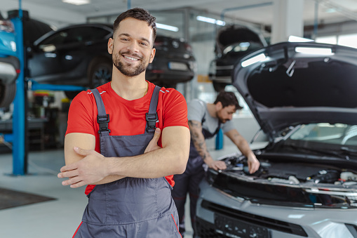 Smiling automotive technician in auto service. The other worker is examining the car