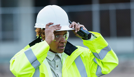 Headshot of an African-American woman working at a construction site, wearing a reflective jacket and safety goggles, putting a hardhat on her head.