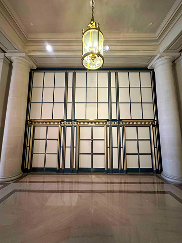 A geometric covering on a marble wall. There is a large lamp hanging from the ceiling in front of the covering. There is a brown marble floor leading up to the covering. This is located in the City Hall, San Francisco.