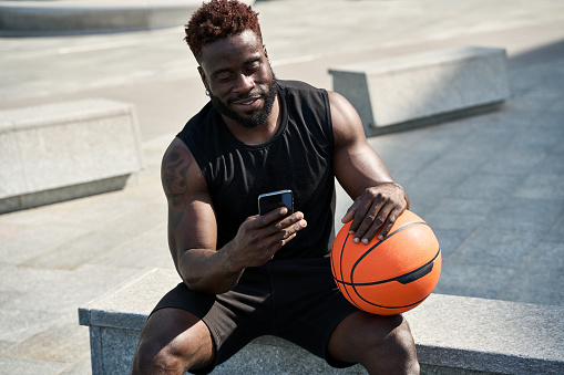 Fit sporty young African black ethnic man sitting outdoors holding basketball ball and mobile phone using apps, looking at smartphone, resting after street sport game with cellphone.