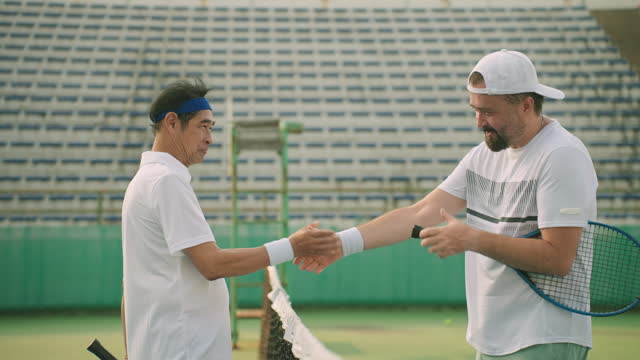 Side view: Caucasian and Chinese senior tennis players shaking hands in front of the net on the court after finishing the tennis-friendly match, Handshake senior athletes, Athlete tournaments, Exercise for good health and well-being