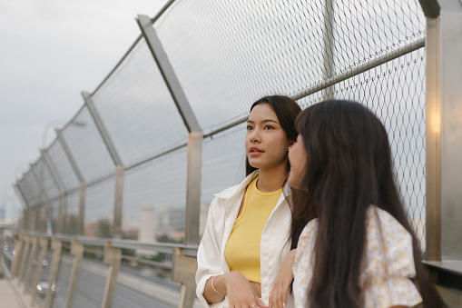 Two young women lean against the chainlink fence talking during the sunrise on a bridge over a chainlink fence