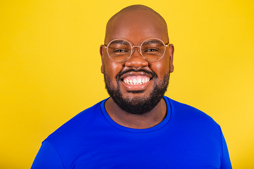 handsome afro brazilian man wearing glasses, blue shirt over yellow background. closeup photo showing smile, concept of happiness, joy, peace, euphoria.