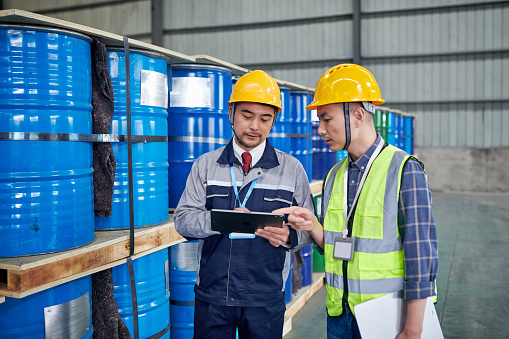 Two asian male worker examining & discussing in chemicals buckets warehouse.