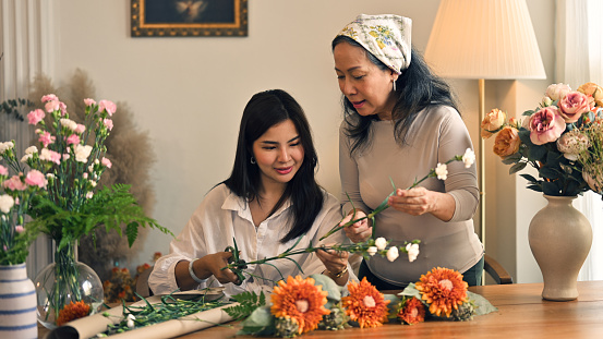 Kind and caring Asian mom teaches her daughter to arrange a vase with beautiful fresh flowers in a living room, spending happy family time on the weekend together.