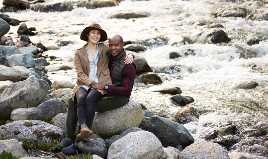 A Japanese woman sits on the lap of an African American man on a large rock by a flowing river in nature. Both looking at camera.