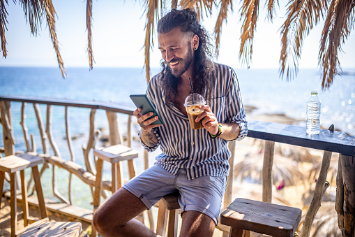 Close-up shot of a handsome young man sitting at the beach bar, using mobile phone and drinking coffee.