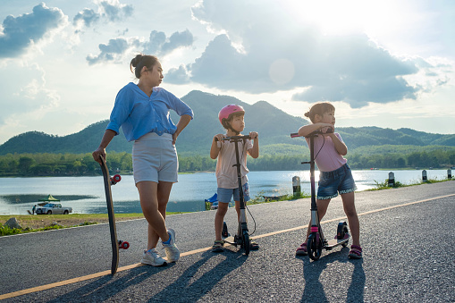 Mother and her children spend their free time playing skateboards and scooters outdoors together. Healthy sports and outdoor activities for school children in the summer.