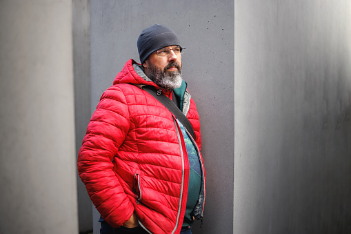 Mid adult bearded man standing outdoors, wearing red winter jacket unzipped and a beanie, size inclusive clothing