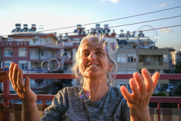 Woman with white hair.Green eyes.Portrait shot on the balcony of the house at sunset.Very cheerful life force.Positive emotion. She plays with her child. Her granddaughter blows bubbles from foam. Grandmother tries to catch her with her palm. Neighborhood buildings in the background. Clear sky.