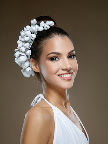 Portrait of beautiful young brunette with brown hair in bun and white fascinator, wearing white sleeveless top and smiling at camera, studio shot, beauty and fashion industry