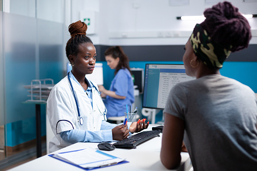 African american doctor talking with woman patient, offering check-up consultation in busy diverse medical workplace. Patient receiving professional examination from consulting general practitioner
