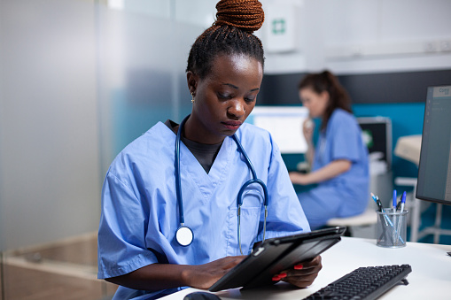 African american nurse working on digital touchscreen tablet in professional healthcare hospital office. Young adult woman medical assistant doing tasks at modern clinic desk, close-up