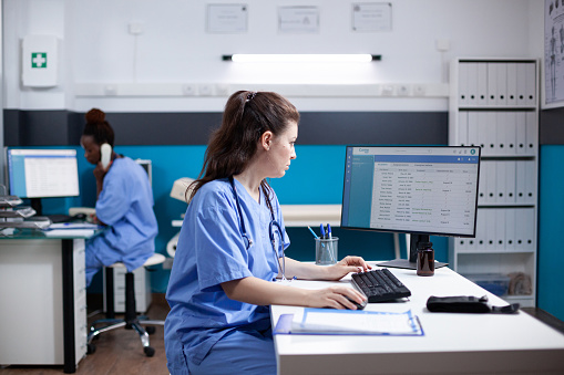 Caucasian nurse sitting at clinic desk, writing data in appointment table list. Focused medical assistant employee wearing scrubs and stethoscope in clean, modern medical office