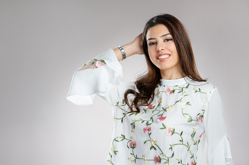 Portrait of beautiful young brunette in white shirt with floral embroidery smiling happily at camera with hand in long hair, studio shot,  beauty and fashion industry