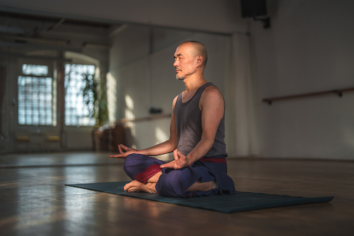 A senior Japanese man meditating in a yoga studio. He sits in a comfortable cross-legged position, with his eyes closed and focused on his breathing.