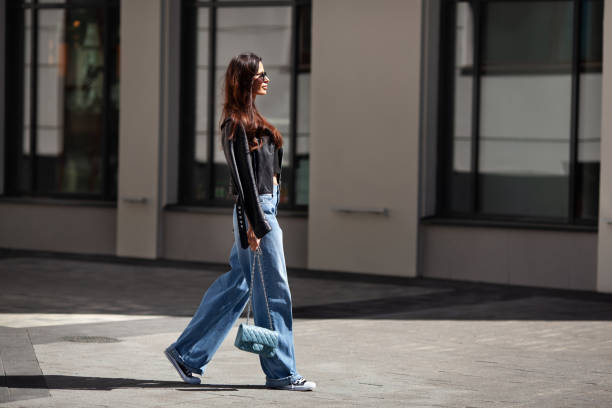 belle belle femme brune vêtue d’une veste en cuir noir et d’un jean bleu large, de baskets, de lunettes de soleil et d’un petit sac à main avec chaîne, rue piétonne par temps ensoleillé - fashion shoe outdoors beautiful photos et images de collection