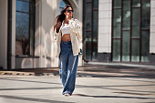 Fashionable woman wearing trendy white bomber jacket, CROP TOP, wide jeans walks on urban city street. Trends clothing of spring and summer