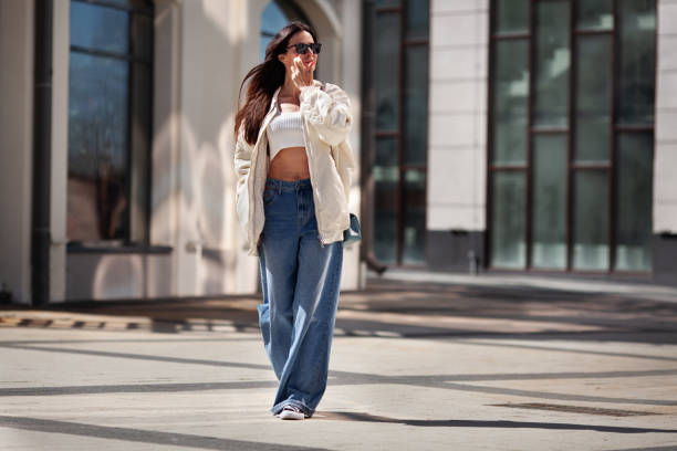 Fashionable woman wearing trendy white bomber jacket, CROP TOP, wide jeans walks on urban city street. Trends clothing of spring and summer Fashionable woman wearing trendy white bomber jacket, CROP TOP, wide jeans walks on urban city street. Trends clothing of spring and summer. flare pants stock pictures, royalty-free photos & images