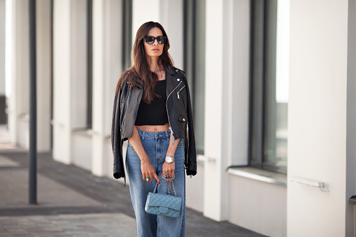 Portrait of gorgeous brunette woman standing city street. Fashion model wears black leather jacket, jeans, short top, small handbag on chain. Street style.