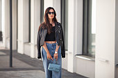 Portrait of gorgeous brunette woman standing city street. Fashion model wears black leather jacket, jeans, short top, small handbag on chain. Street style