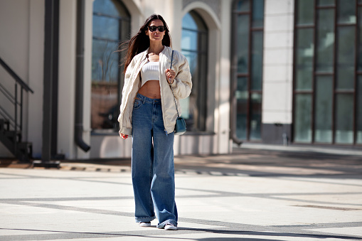 Fashionable woman with long flowing hair wearing trendy white bomber jacket, short Tshirt, wide jeans, posing on street of city. Trends clothing of spring and summer.
