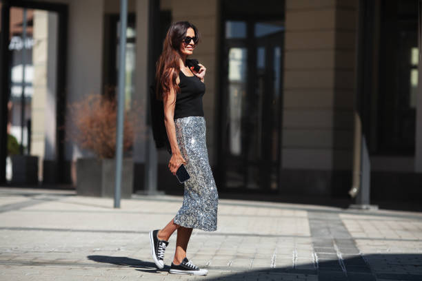 Happy Beautiful brunette woman walks street city near buildings in summer, dressed luxurious clothes shiny maxi skirt with sequins, black sneakers and Tshirt, smiling stock photo