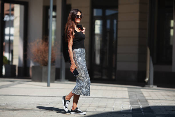 Happy Beautiful brunette woman walks street city near buildings in summer, dressed luxurious clothes shiny maxi skirt with sequins, black sneakers and Tshirt, smiling Happy Beautiful brunette woman walks street city near buildings in summer, dressed luxurious clothes shiny maxi skirt with sequins, black sneakers and Tshirt, smiling. maxi length stock pictures, royalty-free photos & images