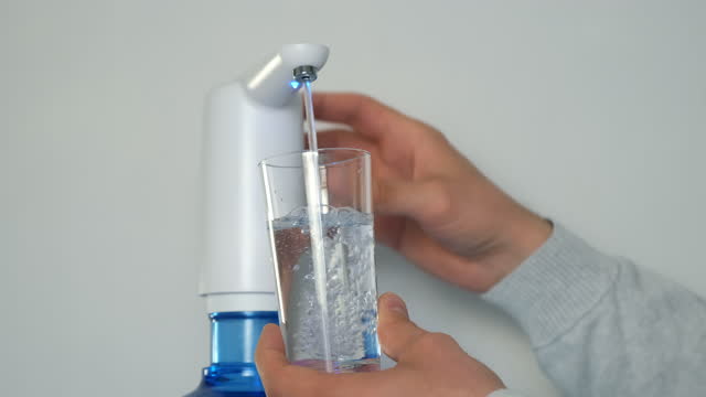 Man's hands pour water into glass from an automatic water cooler, closeup view.
