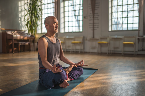 A peaceful moment of a Japanese man in his late 50's meditating with closed eyes on a yoga mat in a bright and spacious studio. The sun shining into the space creates a serene atmosphere that enhances his meditation experience.