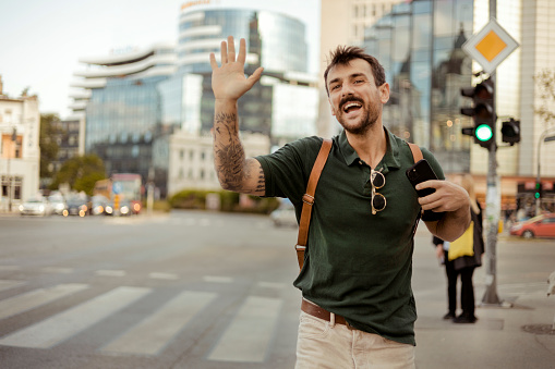 Smiling handsome bearded young man waves joyfully his hand to someone while walking on the city street