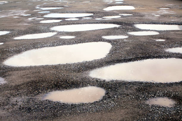 A large pothole filled with water on a rural road reflects the cloudy sky and surrounding trees, while a car speeds past in the background. The image highlights road conditions in a natural, countryside setting.