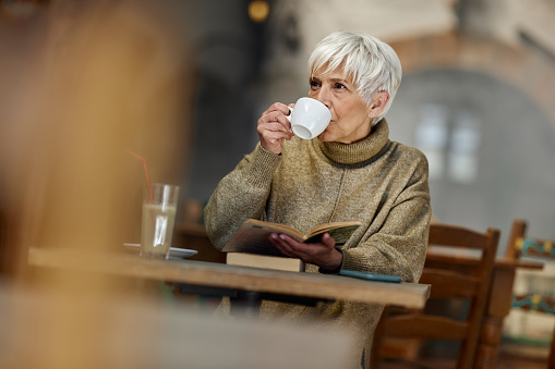 Senior woman drinking coffee while relaxing with a book in a café.
