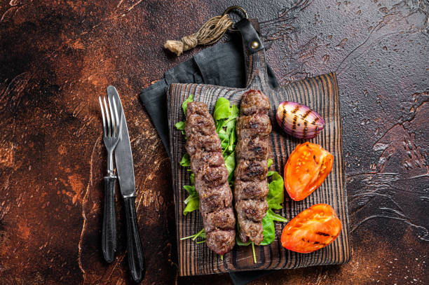 kébab adana turc, bœuf haché et viande d’agneau grillés sur des brochettes servis avec tomate, salade et oignon. fond sombre. vue de dessus - lamb kebab kofta ground photos et images de collection