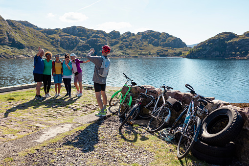 A shot of a small group of senior friends standing by the bay near the village of Diabaig on the side of Loch Torridon in Wester Ross, Scotland. They are taking a group photo on a smartphone and they are enjoying the nice weather. They are on a bike ride.