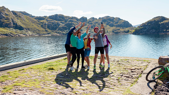 A shot of a small group of senior friends standing by the bay near the village of Diabaig on the side of Loch Torridon in Wester Ross, Scotland. They are taking a selfie on a smartphone and they are enjoying the nice weather. They are on a bike ride.