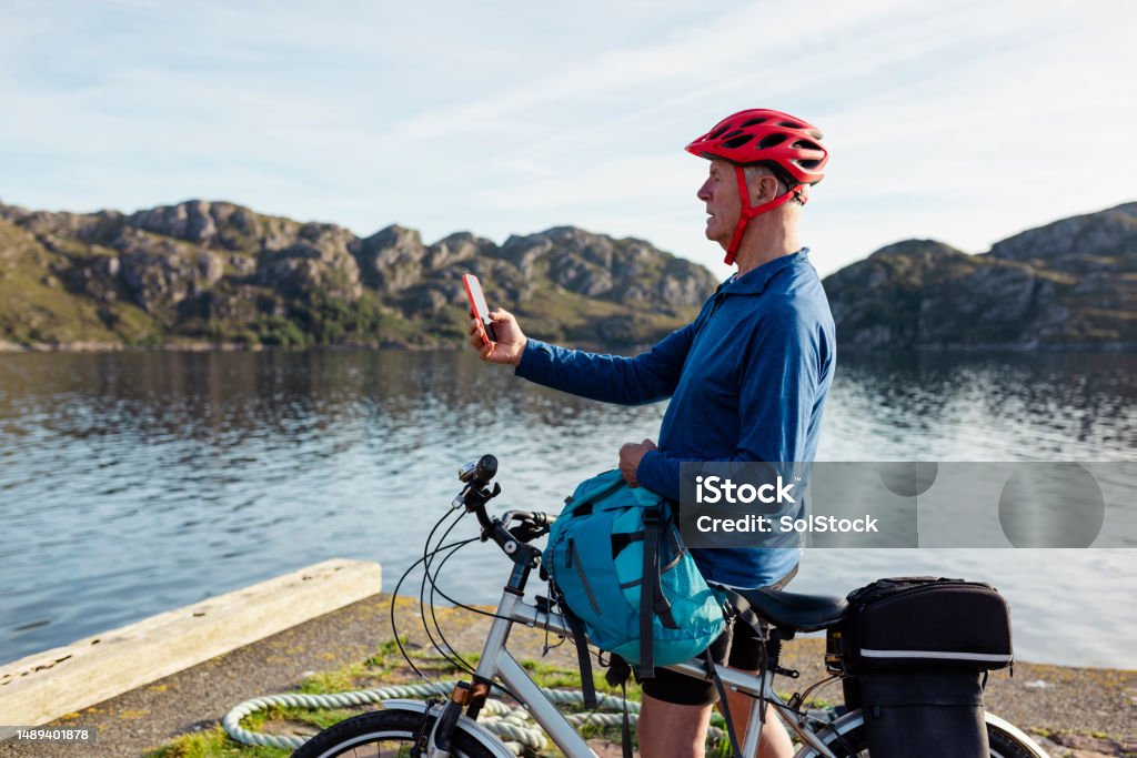Staying Connected on a Rural Bike Ride Senior man standing by the bay near the village of Diabaig on the side of Loch Torridon in Wester Ross, Scotland. He is wearing a bike helmet, on a ride. He has stopped to use his mobile phone. Exercising Stock Photo