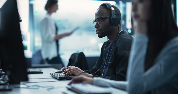 Portrait of a Smart African Young Specialist Working in a Technological Startup Help Desk Department, Using Computer in a Diverse Office. Black Man Wearing Glasses and Headphones with a Microphone