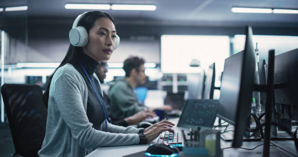 istock Portrait of a Thoughtful Young Engineer Working on Computer in a Technological Office Environment. Beautiful Asian Woman Wearing Headphones, Writing Software Code for a Blockchain Project 1489403965