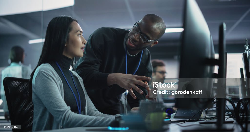 Two Diverse Multiethnic Colleagues Having a Conversation While Busy Working on a Team Project. Asian Female Designer Talking with an African Project Manager. Teamwork in Technology Laboratory Artificial Intelligence Stock Photo