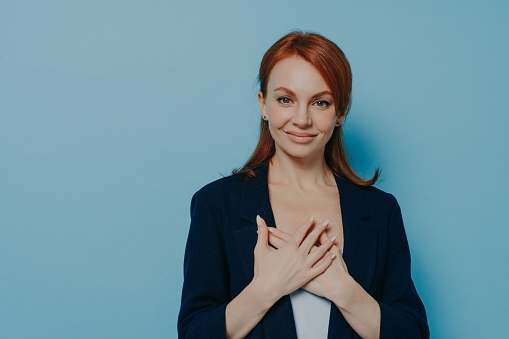 Studio shot of happy smiling woman with red hair holding hands on heart and feeling grateful, thankful, showing gesture of gratitude while standing isolated on blue background, copy space for text