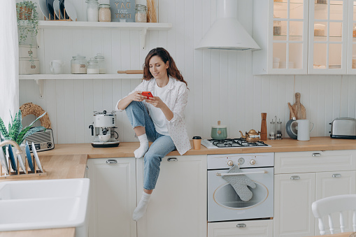 Caucasian young woman has message on cellphone. Happy girl with telephone sitting on worktop of table at kitchen and texting. Modern luxurious scandinavian interior. Lifestyle concept.
