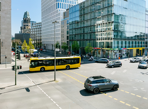 A bird's-eye view of a busy street in Berlin's Mitte district. Cars and public transportation move along the wide street, while pedestrians and cyclists weave in and out of traffic. Office buildings and shops line the street, and the iconic Kaiser-Wilhelm-Gedächtniskirche can be seen in the background. The sun is shining brightly and the sky is a clear blue.