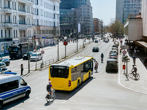 View from above of a busy street in Berlin's Mitte district. Cars and public transportation move along the wide street. Office buildings and various shops line the left and right sides of the street. Pedestrians are visible on the right side of the street.