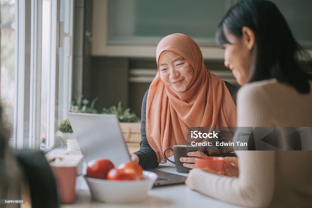 Asian Malay woman in hijab with vitiligo skin condition at kitchen using laptop talking to her female friend Islam Stock Photo